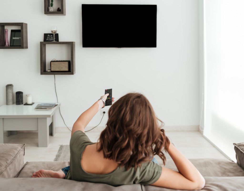 vista-posterior-mujer-casual-sentada-sofa-viendo-television-casa